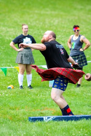 Highland games athlete in tartan kilt demonstrates throwing technique on grass field.