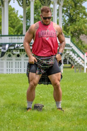 Athletic figure in red tank top and plaid kilt performs a dynamic workout movement on grassy lawn with gazebo backdrop.