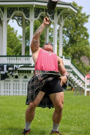 Person in pink tank top and plaid kilt demonstrates athletic stance on sunny lawn near white gazebo.
