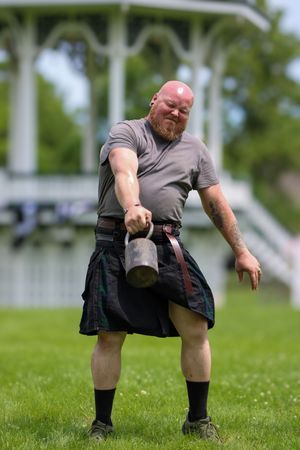 Figure in grey shirt and dark kilt performs strength training exercises on grass with gazebo in background.