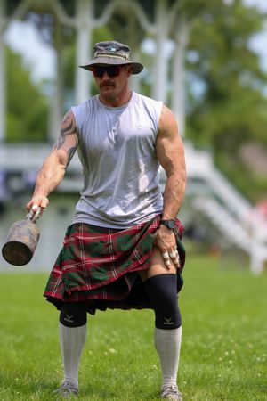 Athletic person in white tank top and plaid kilt demonstrates squatting position during outdoor workout.