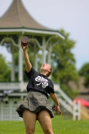 Person in black shirt and plaid skirt performs athletic movement on grass with white gazebo in background.