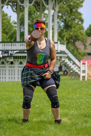Athlete in dark top and plaid kilt demonstrates proper workout form on sunny lawn near gazebo.