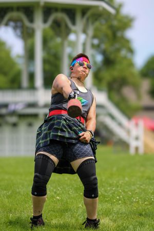 Person in athletic wear and plaid kilt performs training exercises on grass with white gazebo visible.