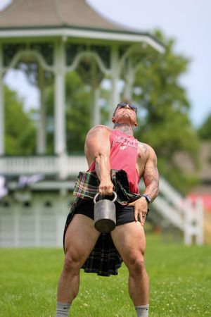 Athlete in red tank top and plaid kilt performs strength training exercise on sunny lawn.