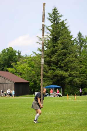 Athletes compete in a caber toss event on a grassy field with spectators and tents visible in the background.