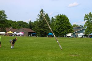 Panoramic view of a caber toss competition with spectator tents and trees in the background.