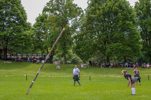 Spectators watch from the sidelines as athletes prepare for caber toss competition on green field.