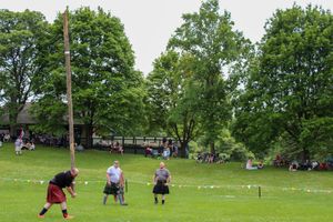 Athletes compete in traditional Scottish Highland games on a grassy field with colorful banners.