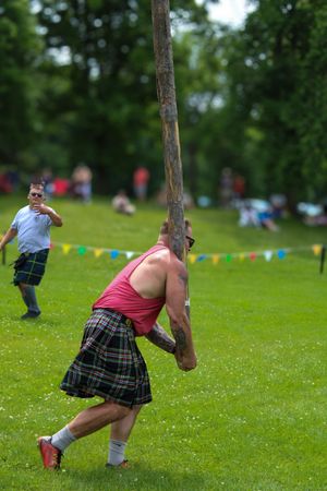 Athlete in traditional Scottish attire performs caber toss on grass field.