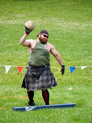 Highland Games athlete in kilt executes hammer throw technique during competition.