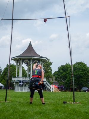 Highland Games athlete demonstrates hammer throw technique on grassy field with gazebo backdrop.