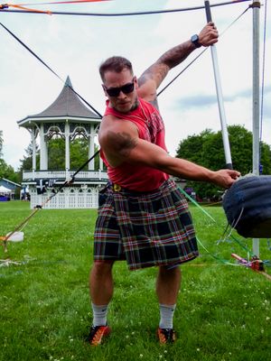 Athletes in traditional Scottish dress compete in throwing events near white gazebo.