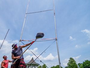 Athletes compete in a Highland Games weight throw event on a sunny day at a grassy field.