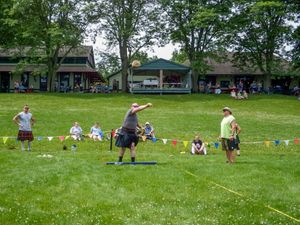 Spectators watch Highland Games athletes compete on a grassy field with buildings in background.
