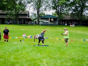 Multiple competitors practice traditional Highland Games events on a green field with spectators watching.
