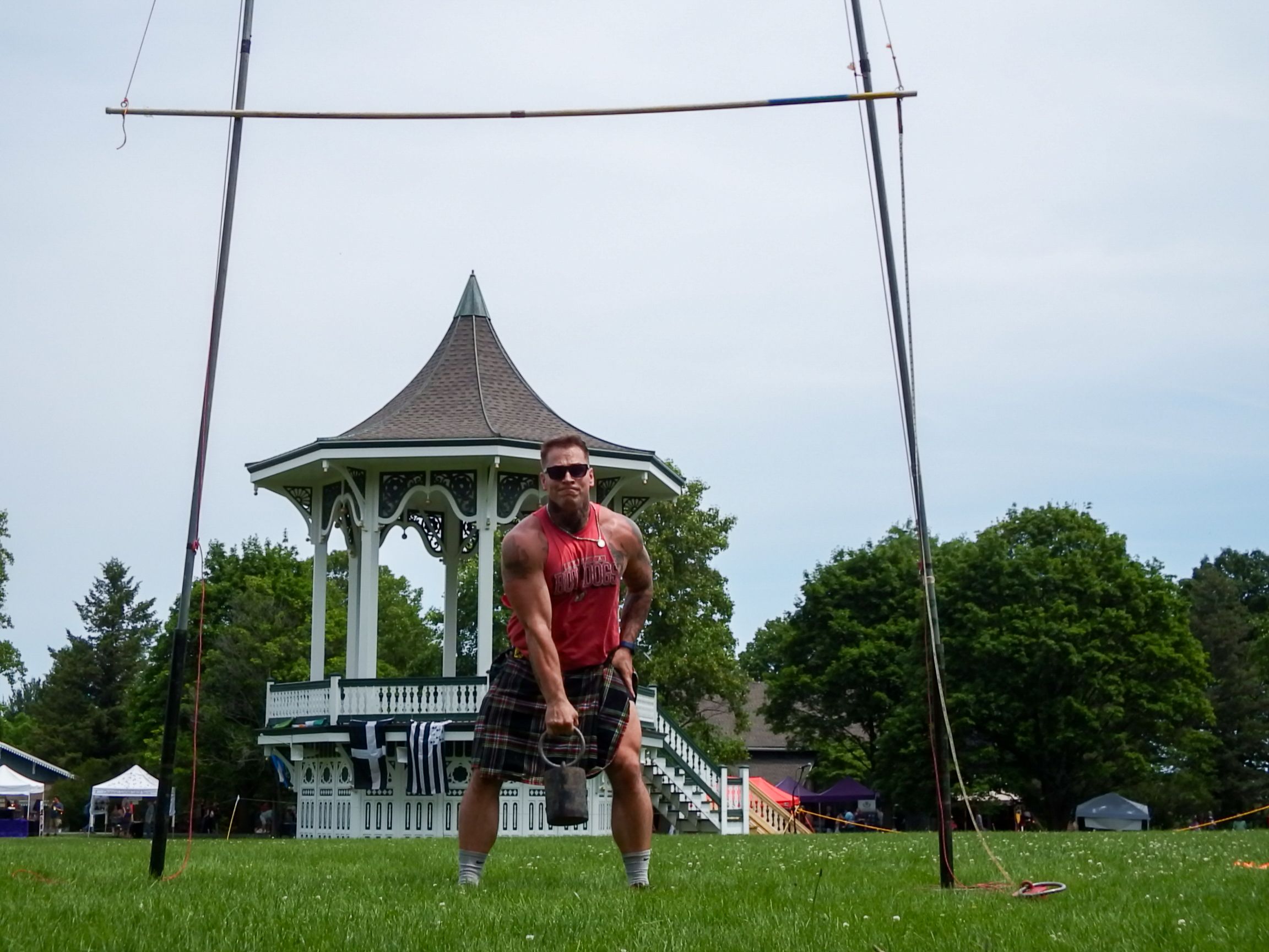 A competitor prepares for a weight throw under a height bar with a gazebo in the background.