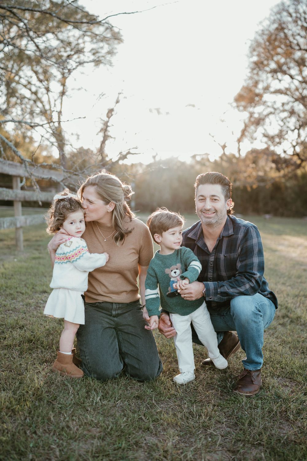 A happy family of four poses outdoors in golden hour light, with parents kneeling beside two young children on green grass.