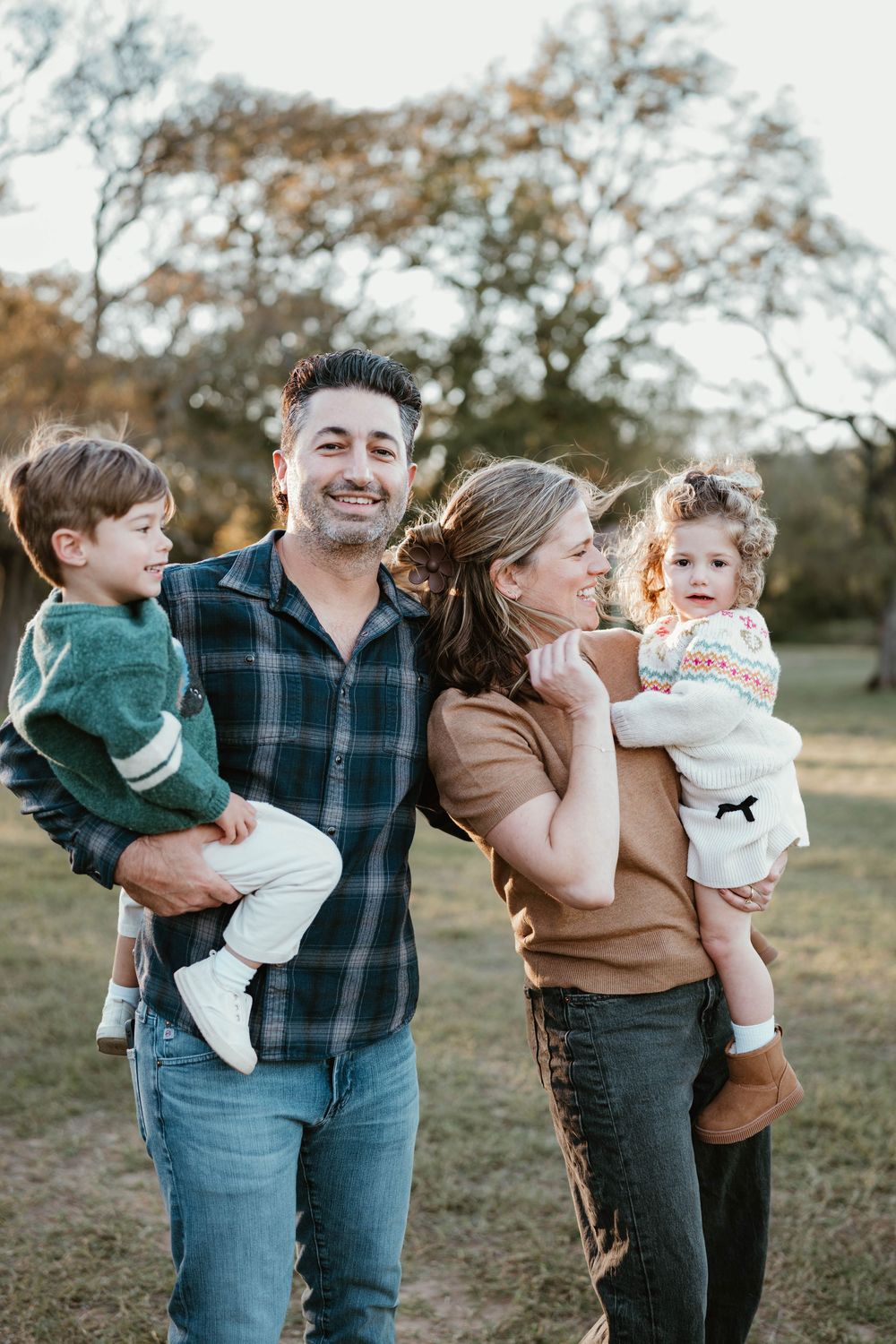 A happy family of four poses outdoors in a park, parents holding two young children, wearing casual fall clothing.