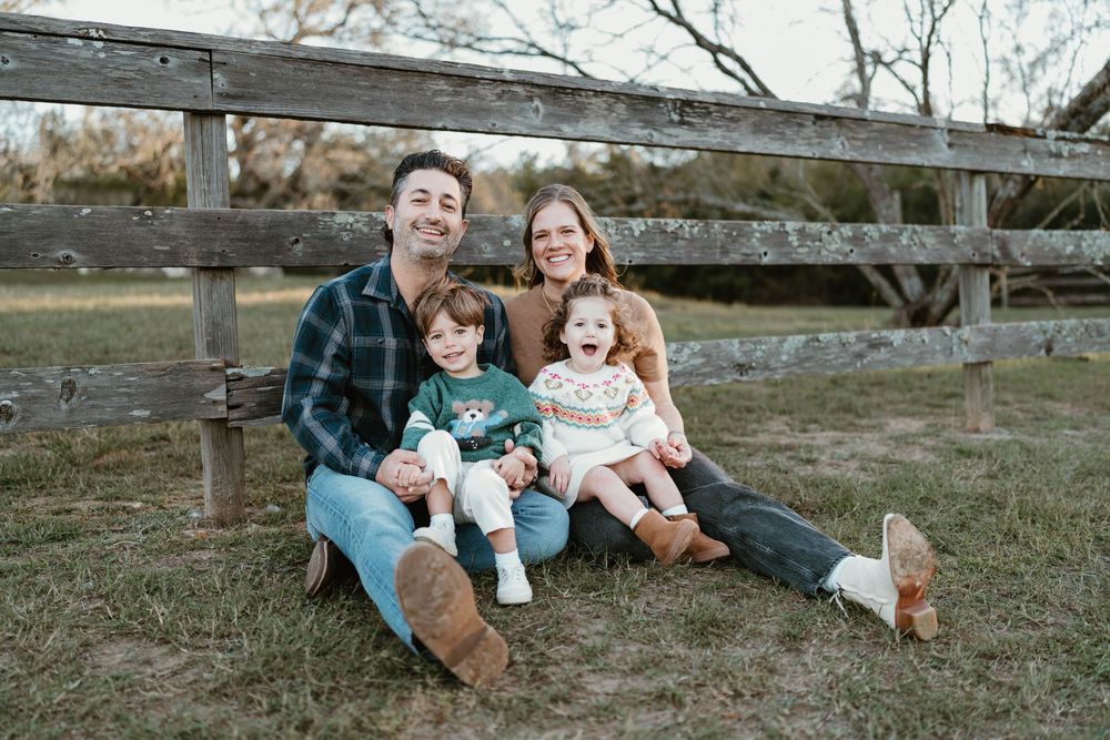 Happy family of four sitting on grass near wooden fence, parents smiling with two young children outdoors.