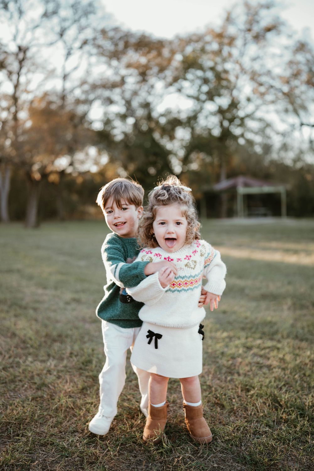 Two young siblings hugging and laughing outdoors in a sunny park, dressed in coordinating fall outfits.
