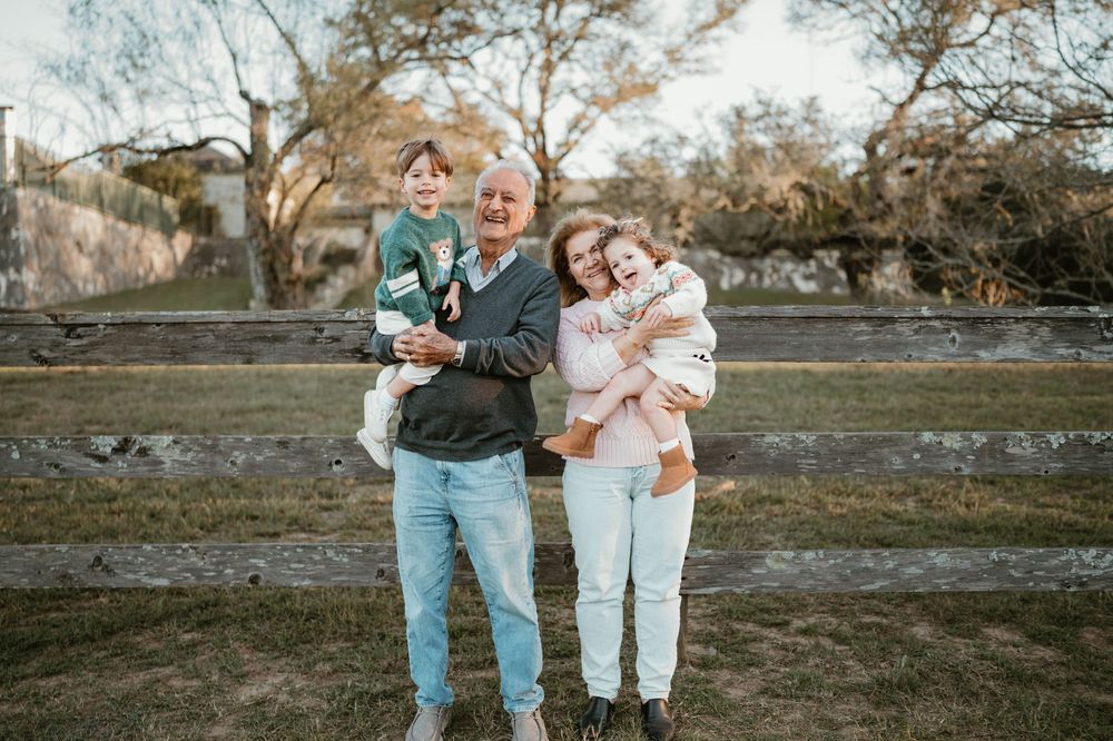 Grandparent smiling outdoors holding two young grandchildren near a wooden fence on a sunny day.