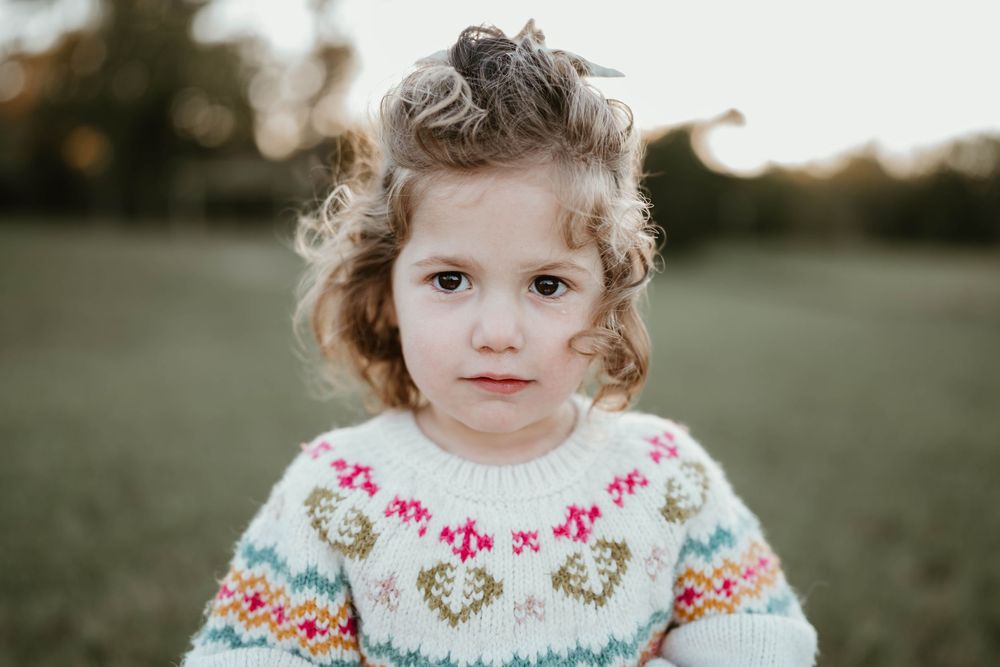 A cute toddler girl with curly hair wearing a colorful heart-patterned knit sweater stands in an outdoor field.