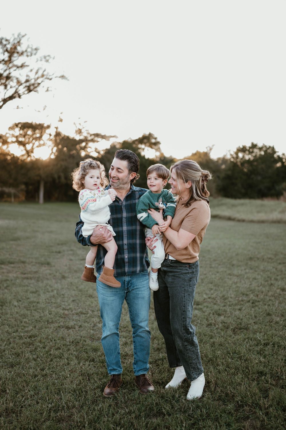 Family of four poses in a sunlit field, parents holding two young children, smiling warmly at golden hour.