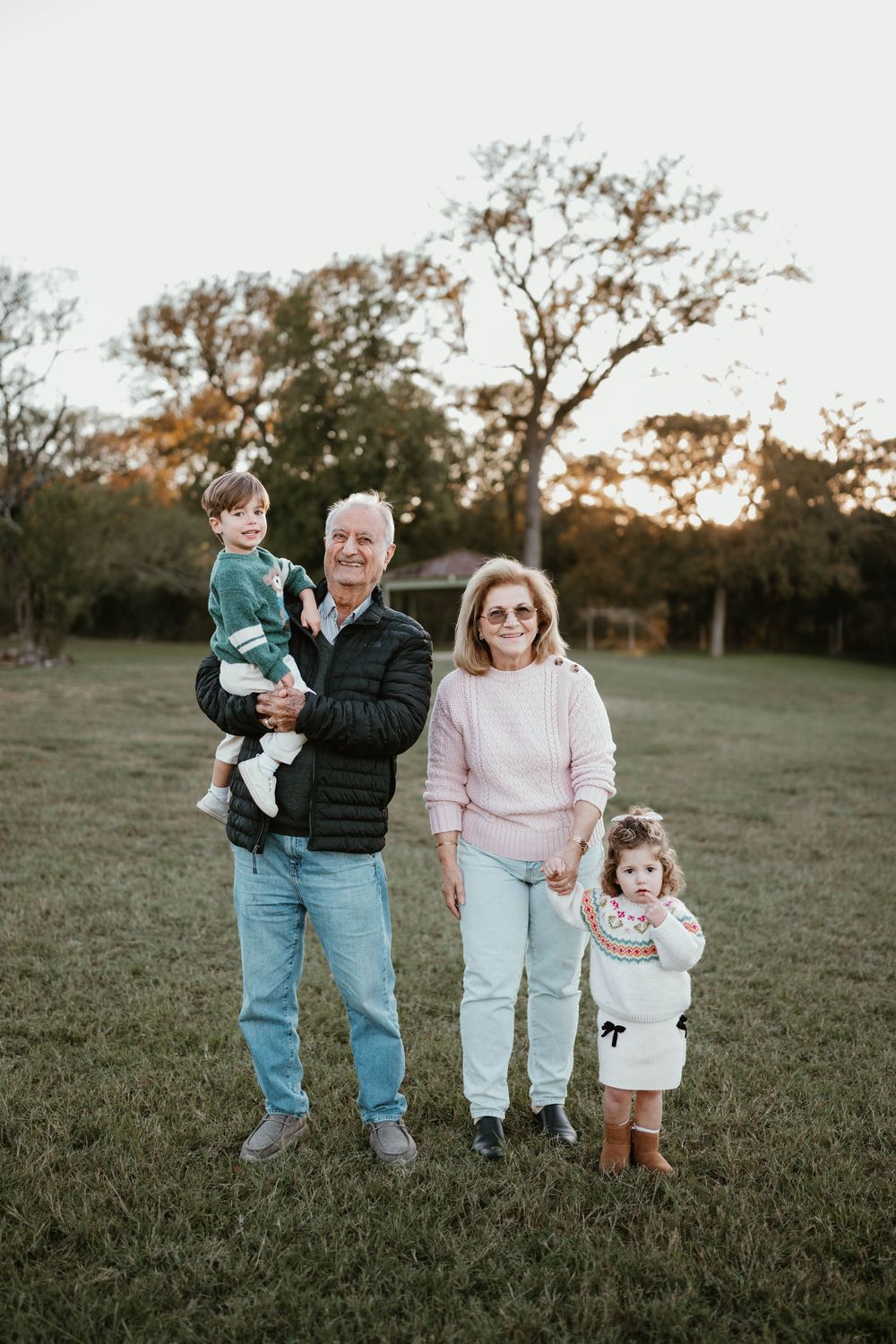 Grandparents posing outdoors with two young grandchildren on a grassy field at sunset.