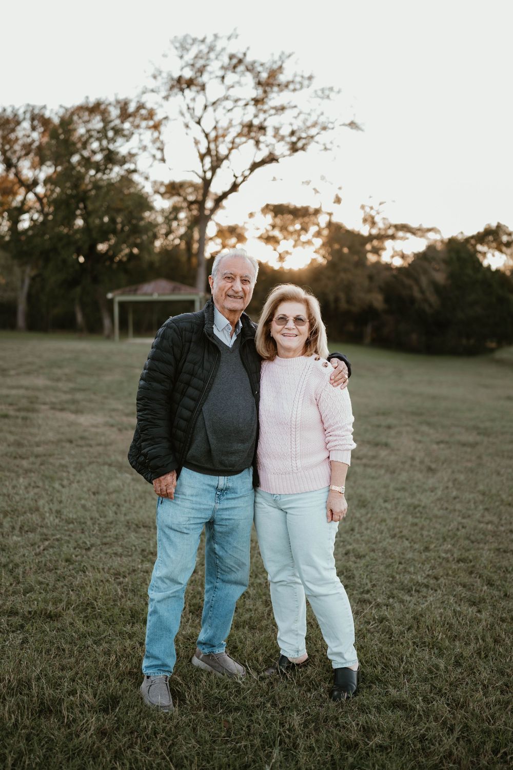 Elderly couple smiling and embracing outdoors on green grass at sunset with trees in background.