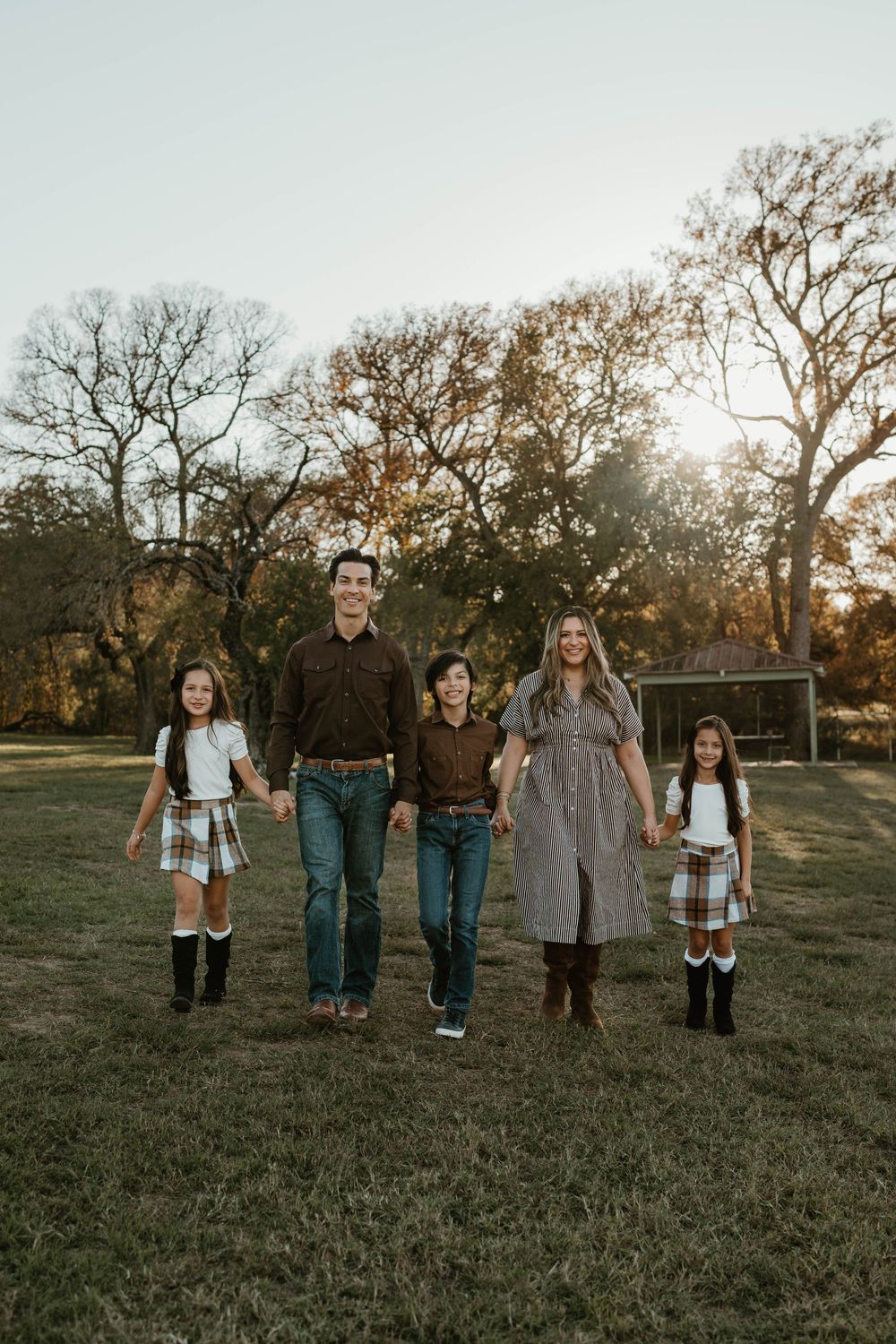 Family of five holding hands walking through a sunlit park with bare oak trees in the background.