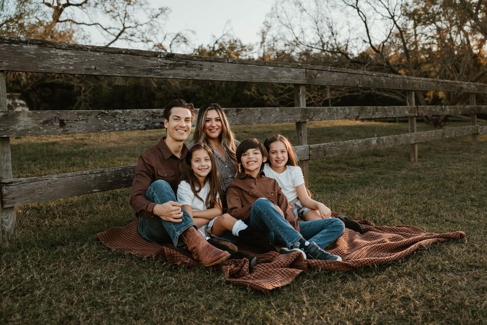 Family of five smiling together on a blanket outdoors by a wooden fence during a fall family portrait session.