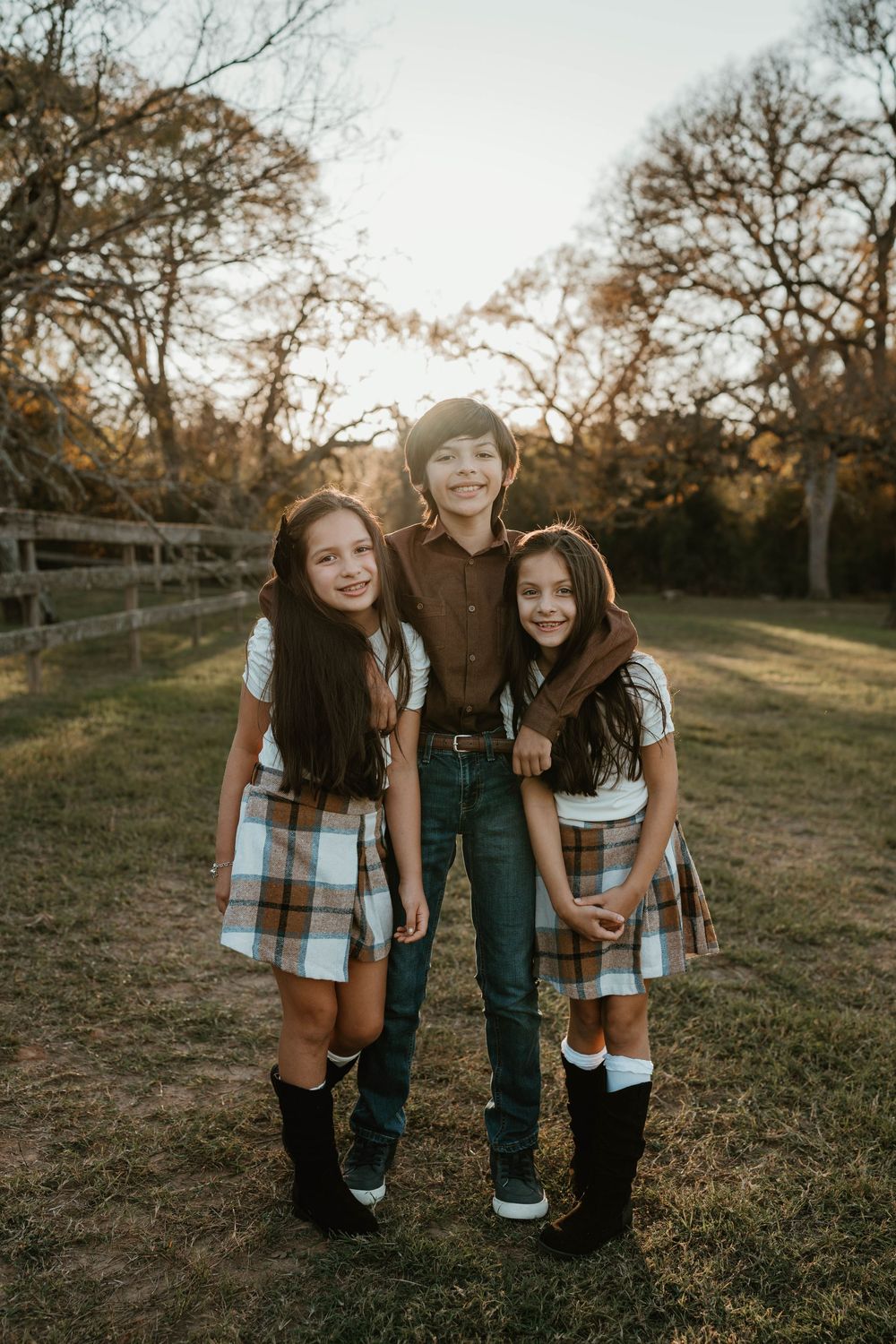 Three children pose outdoors at golden hour, two girls in matching plaid dresses with boots and a boy in jeans and brown shirt.