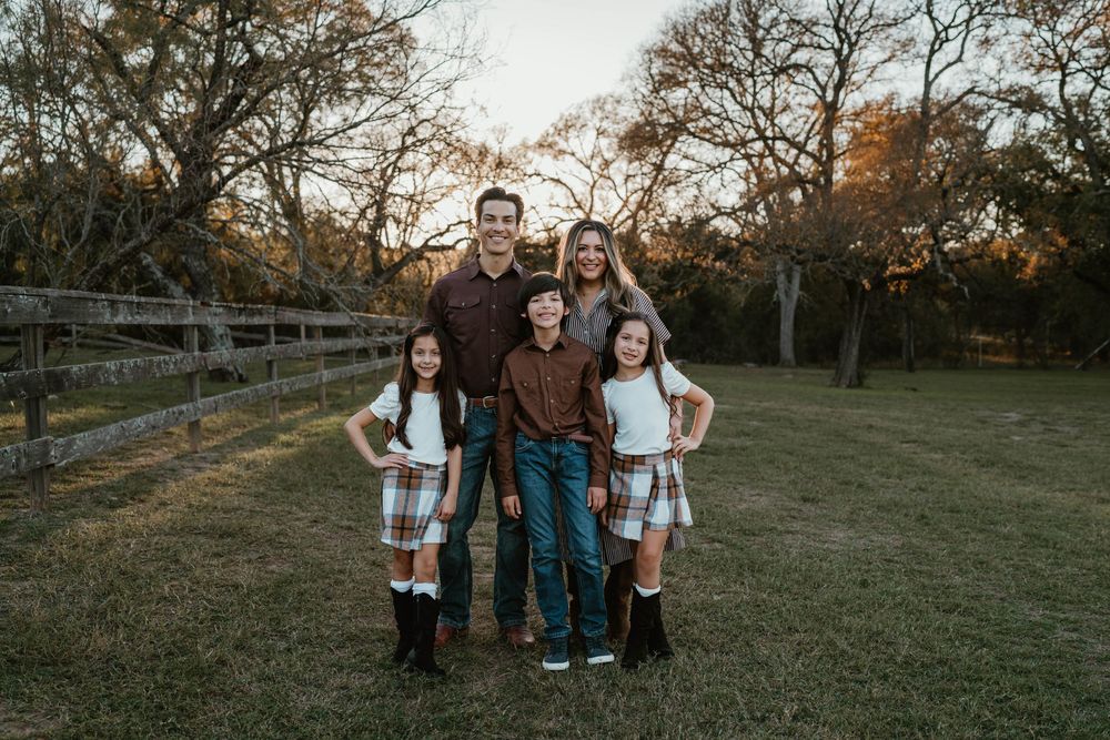 Family of five posing outdoors near a wooden fence with autumn trees in the background during golden hour.