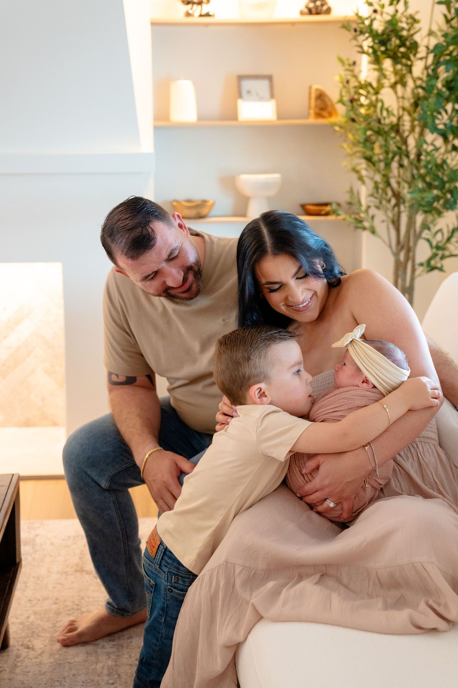 A happy family of four admires their newborn baby together in a cozy, warmly lit living room.