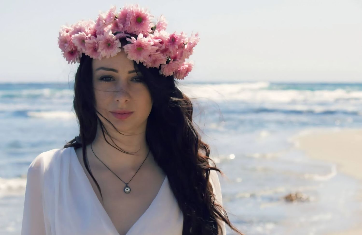 A young woman wearing a crown of flowers poses for a photo while standing near the ocean.