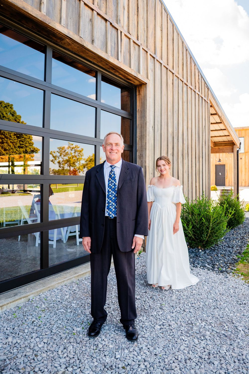 Bride in white gown poses with older man in dark suit outside a rustic barn venue.