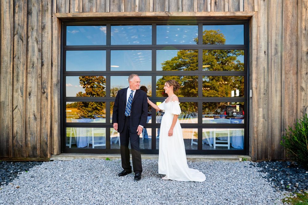 Bride and groom holding hands outside rustic barn wedding venue with large glass doors and gravel path.