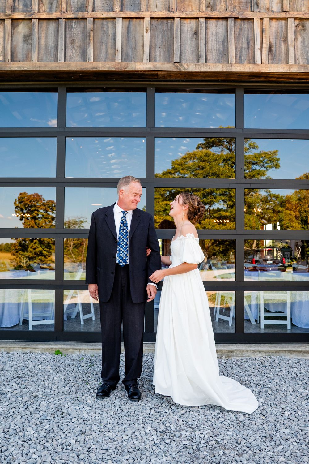 Bride in white off-shoulder gown smiles at older man in suit outside rustic barn venue.