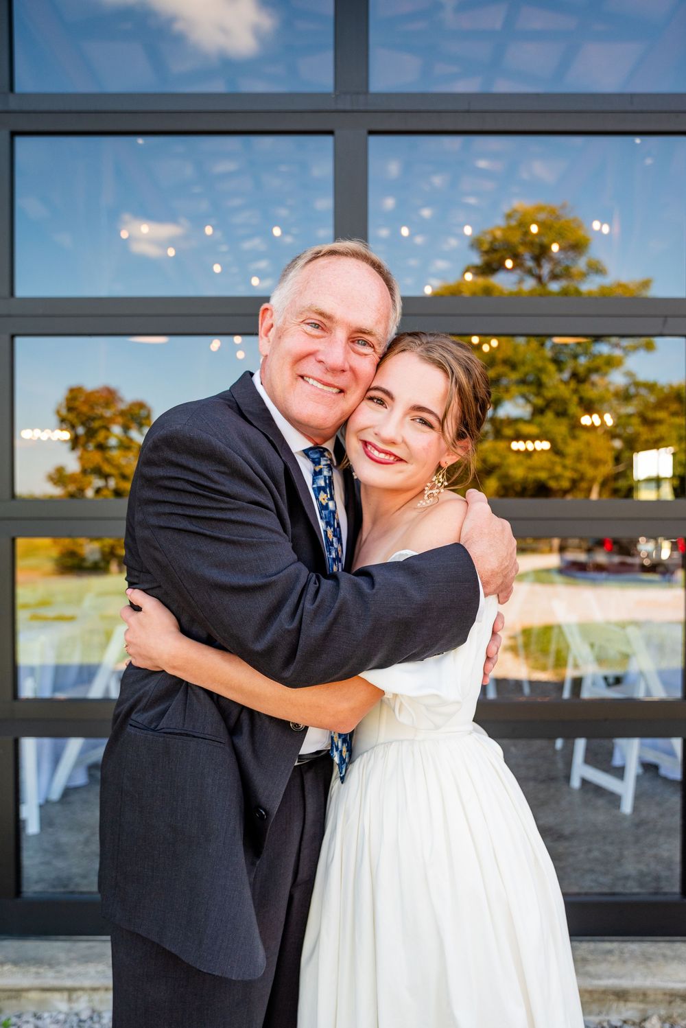 Bride and older man share a warm hug in front of large windows at wedding reception.