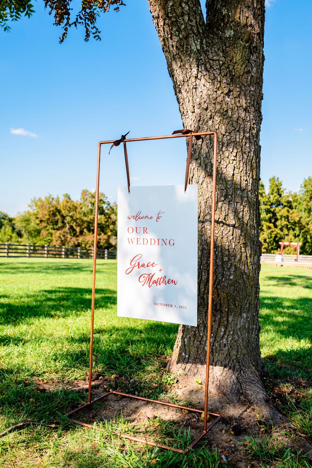 Outdoor wedding welcome sign for Grace and Matthew displayed on copper frame stand near tree.