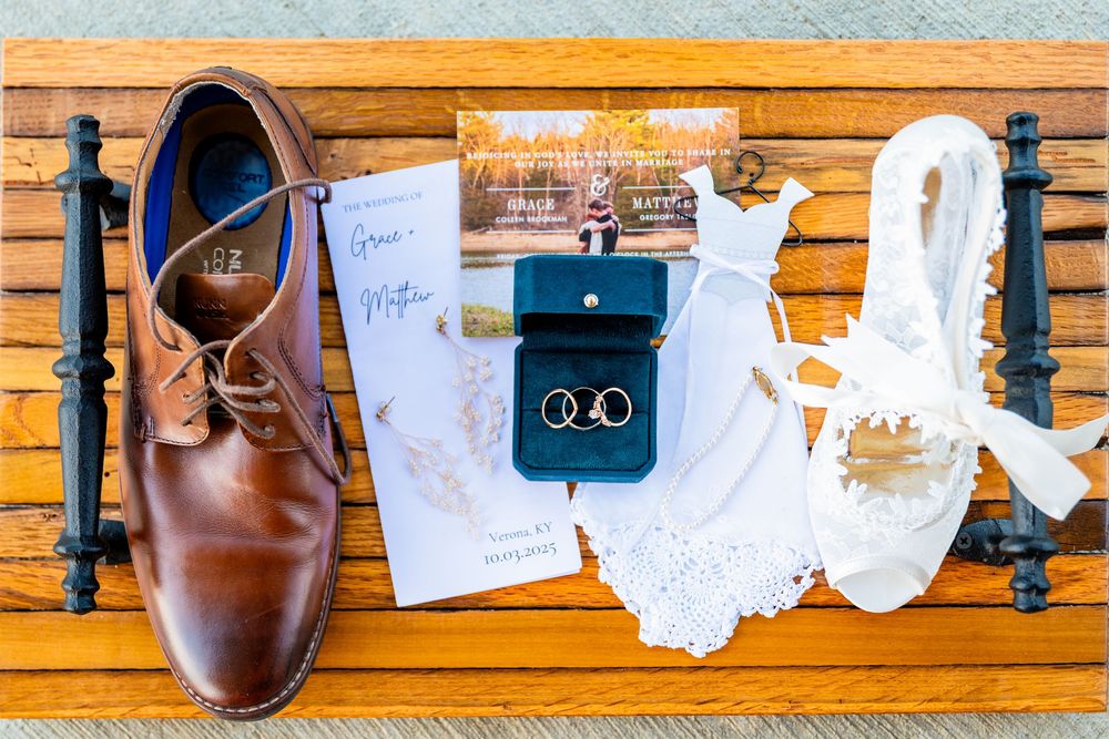 Wedding flat lay with brown dress shoes, rings, invitation, and lace details on bench.