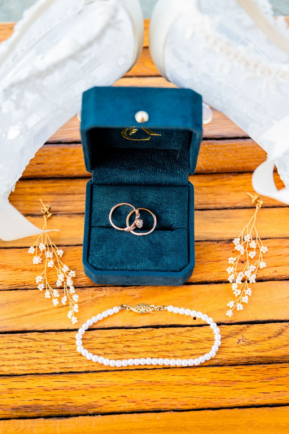 Wedding rings in a teal velvet box with pearl bracelet and floral hair pins on wooden table.