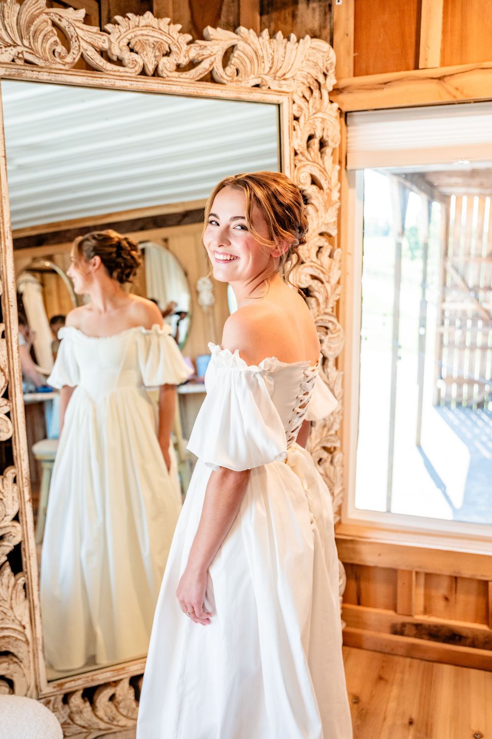 Smiling bride in an off-shoulder white wedding gown posing in front of an ornate carved mirror in a rustic barn.