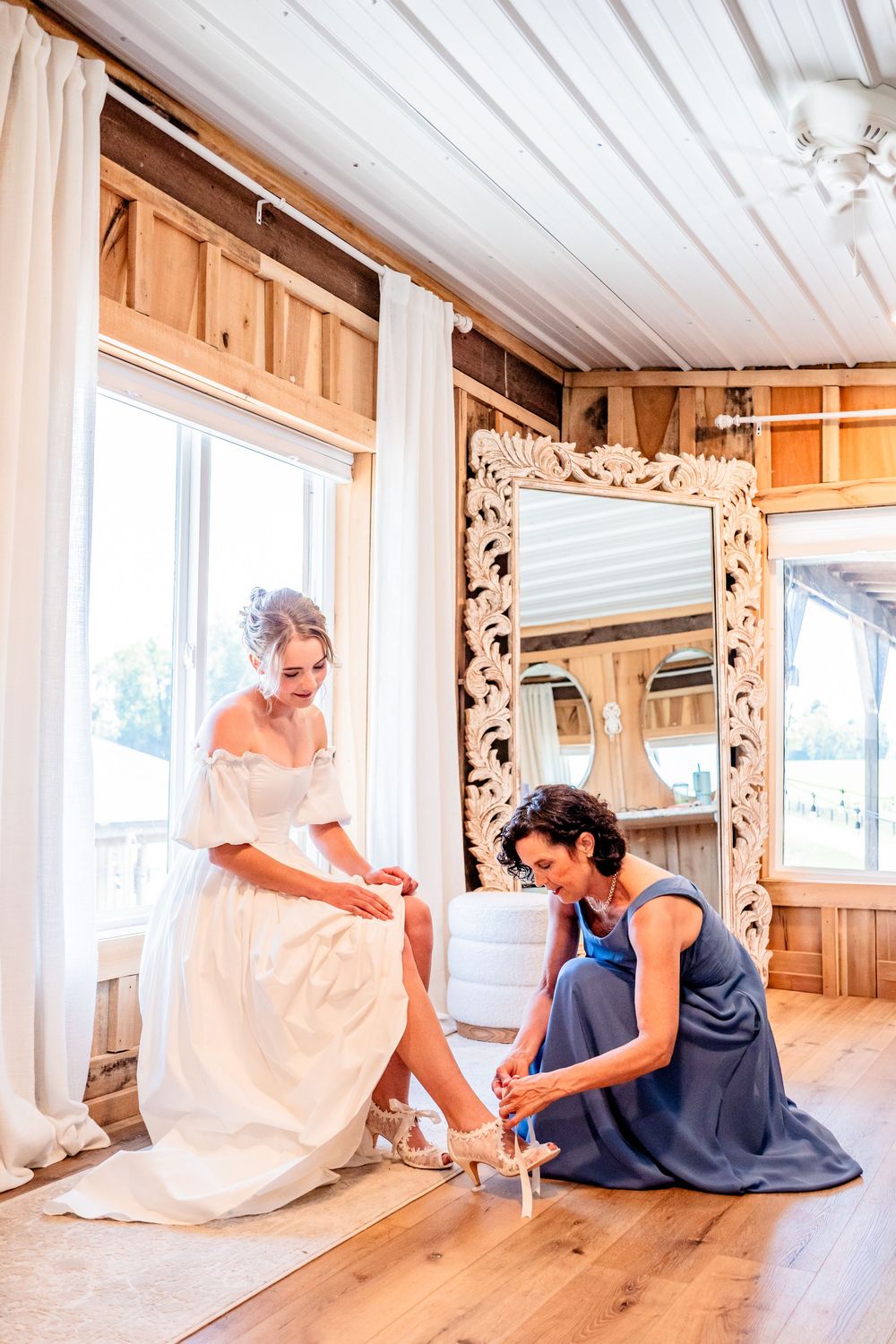 Bridesmaid helping bride put on heels in rustic wooden bridal suite with ornate mirror.
