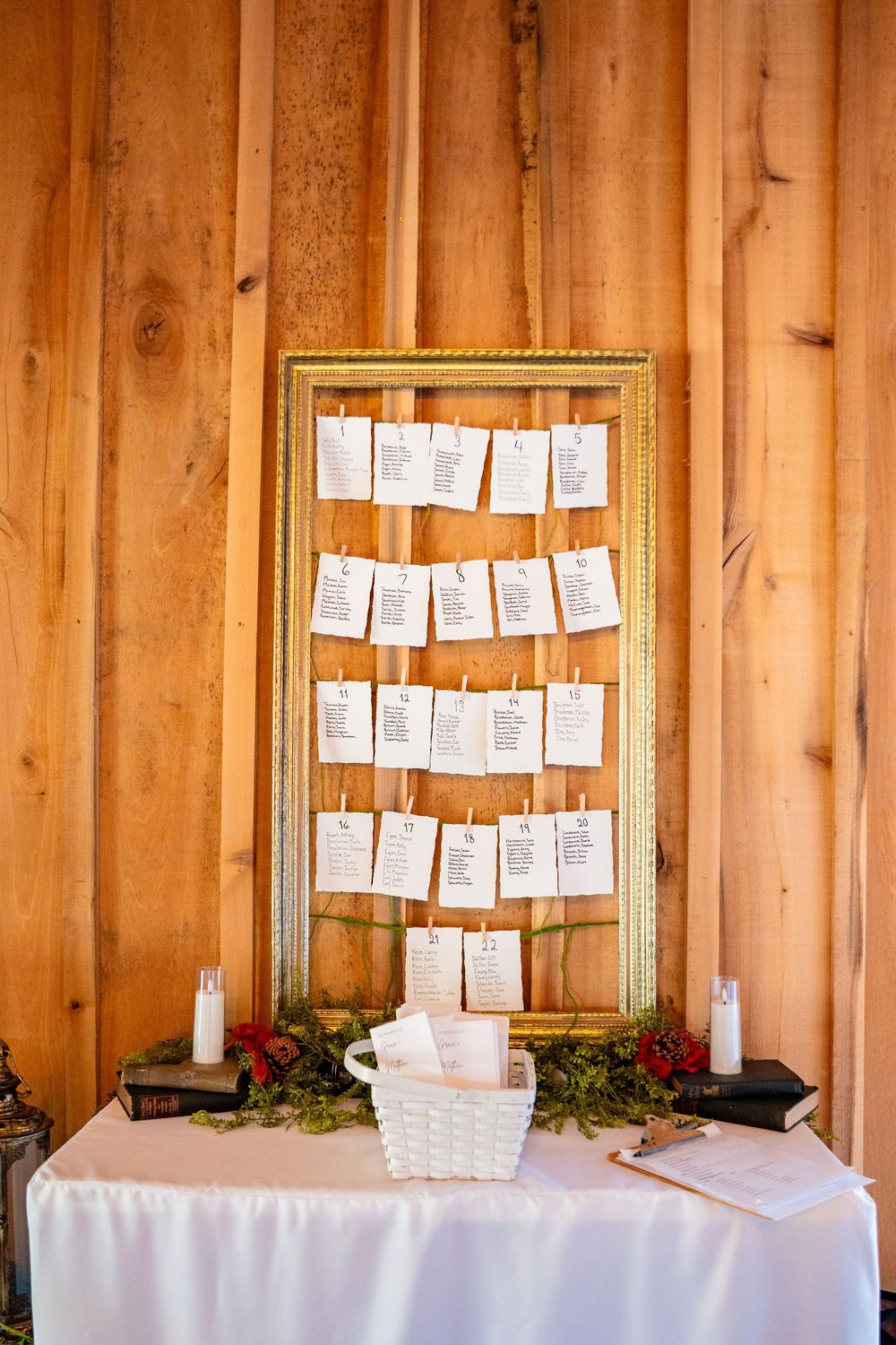 Rustic wedding seating chart displayed on a gold frame with hanging cards on a white draped table in a wood-paneled barn.