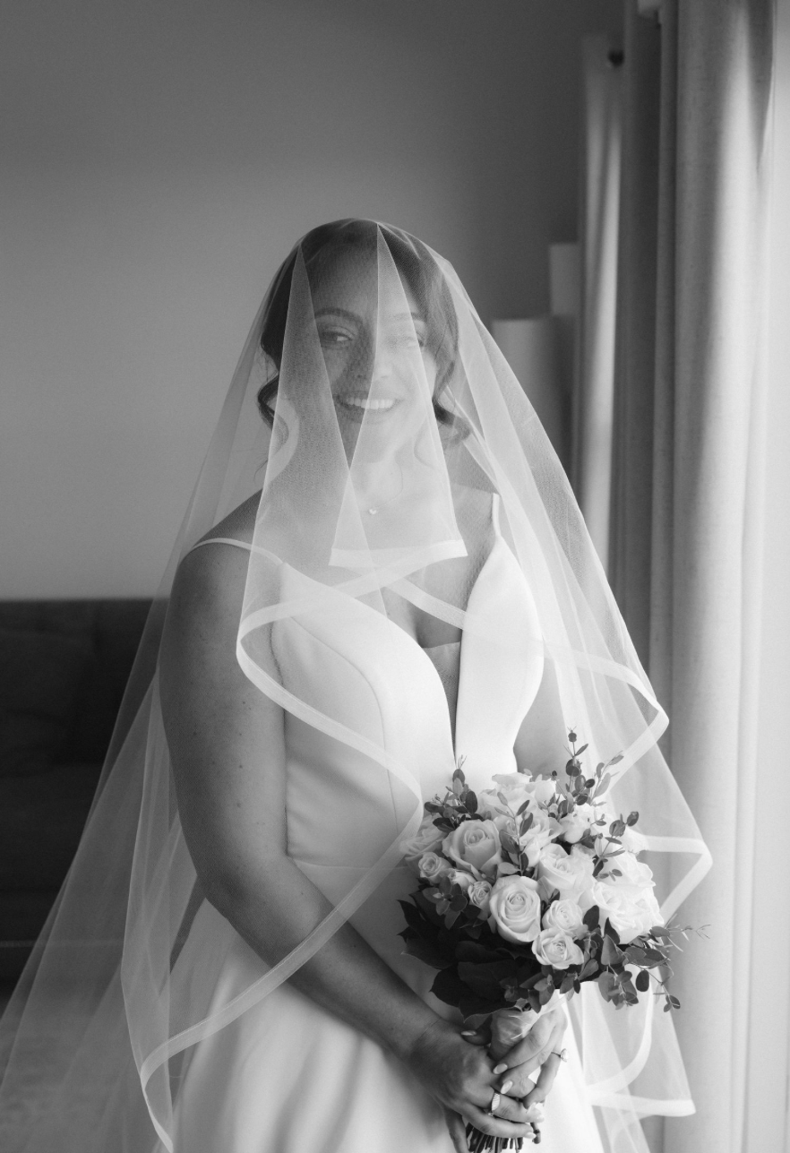 Black and white bridal portrait of smiling bride in strapless gown holding floral bouquet with flowing veil draped over shoulders.