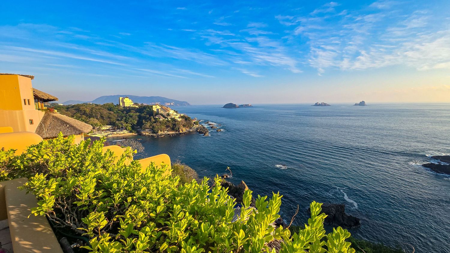 Scenic coastal view with lush green plants, rocky cliffs, and calm blue sea under a bright sky.