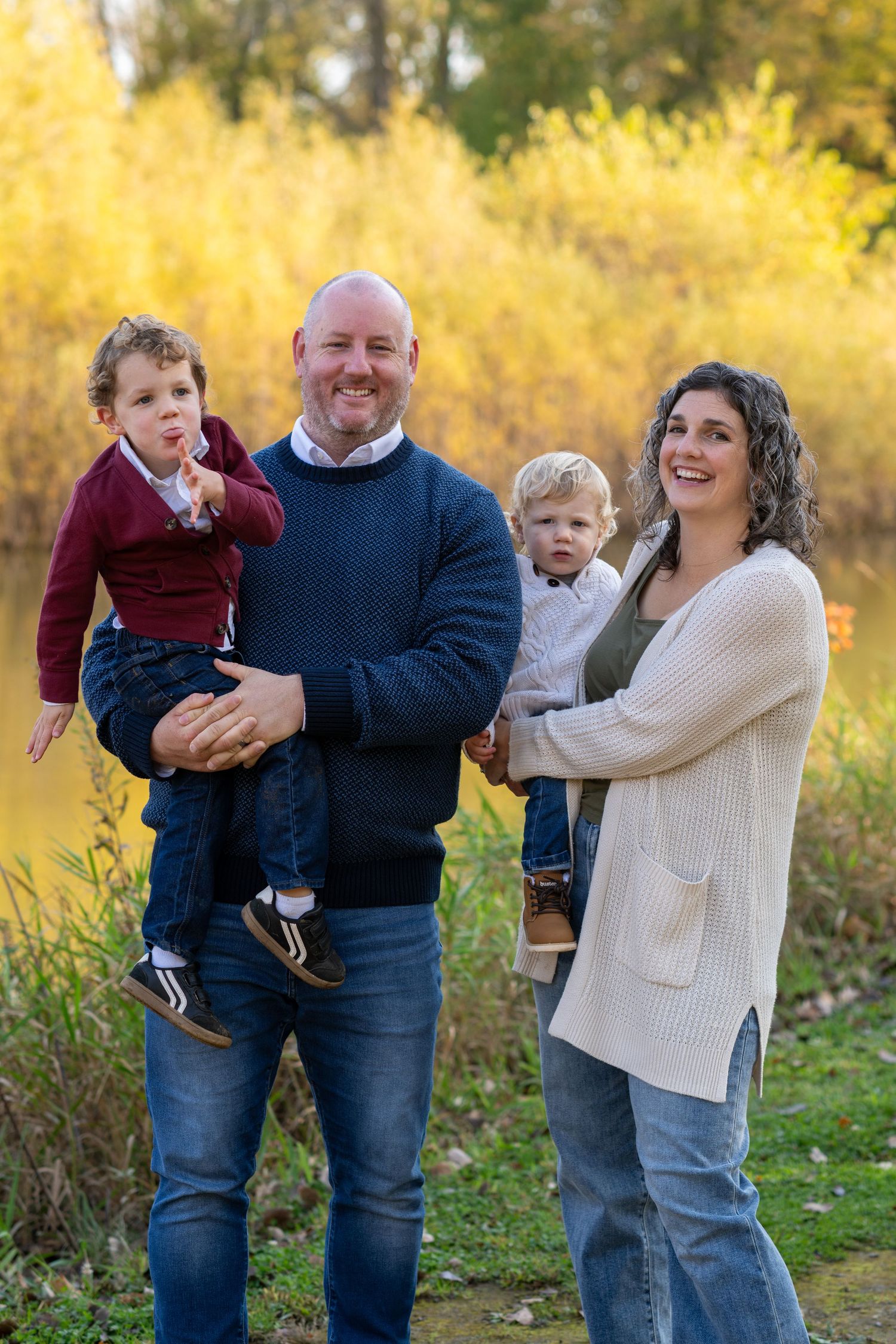 Family of four posing outdoors in fall, parents holding two young boys near a pond with yellow foliage.