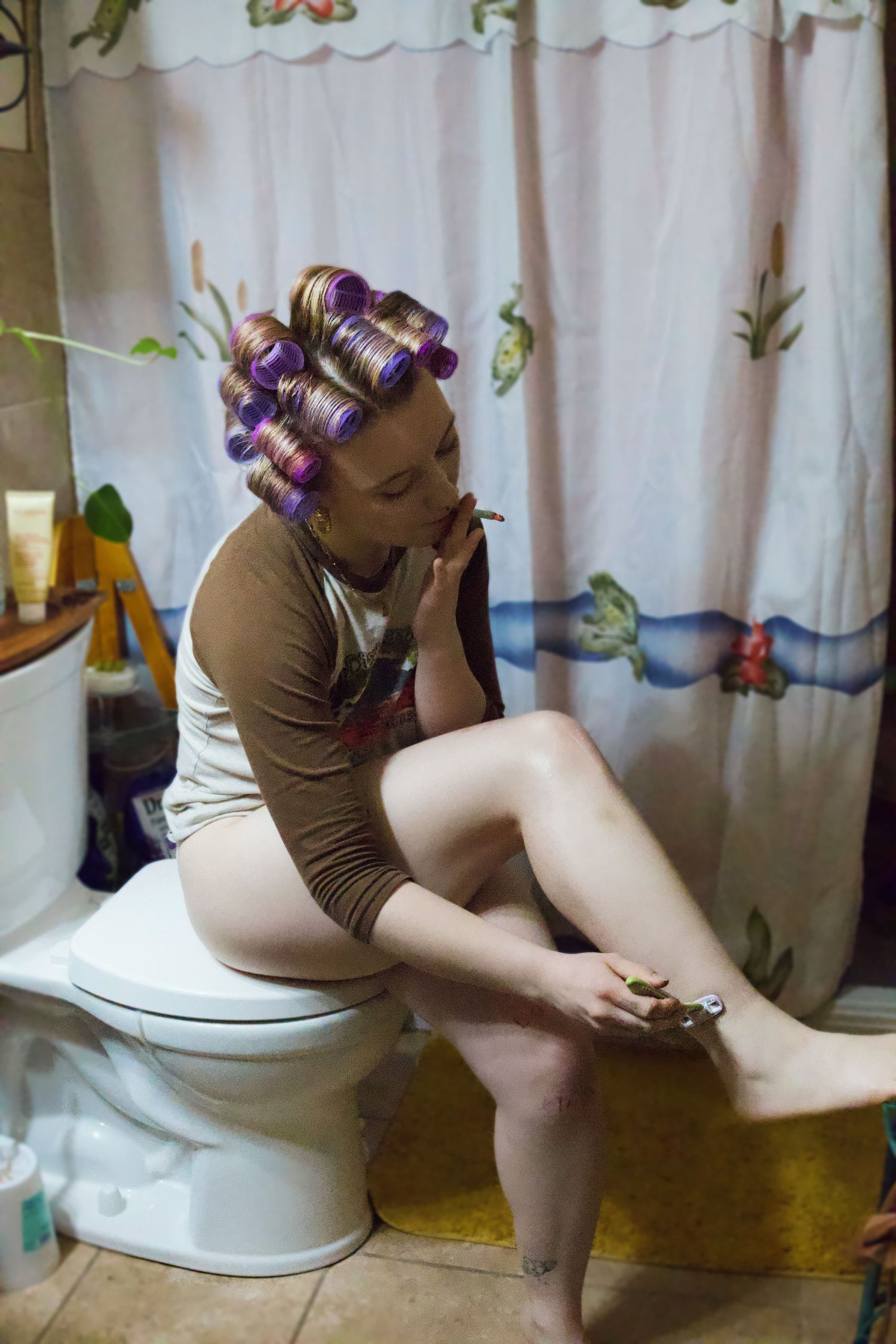 Woman with purple hair rollers sits on toilet edge in floral bathroom, shaving legs near Camel cigarettes.